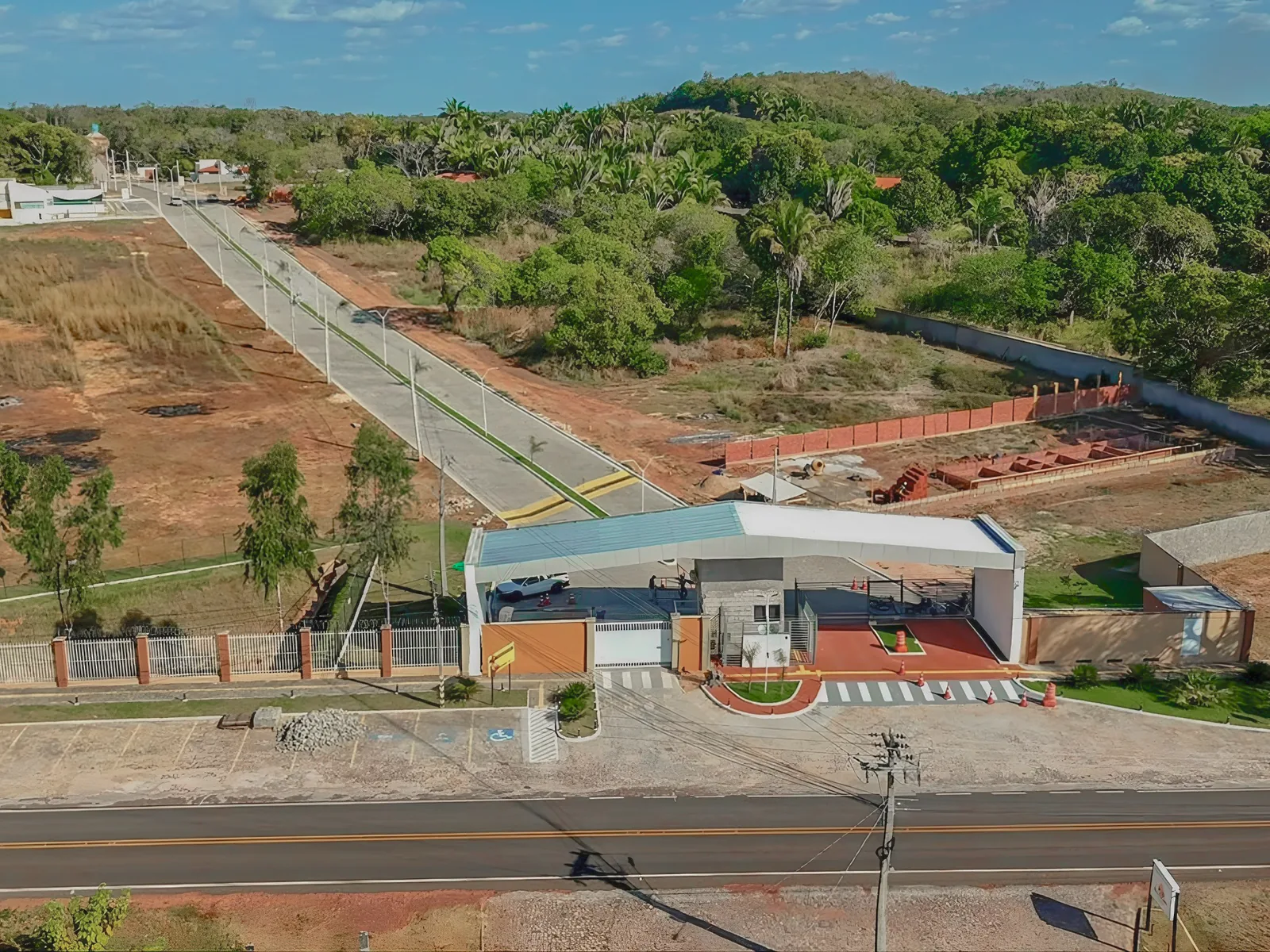 Vista aérea da entrada do condomínio com via asfaltada, iluminação pública e muro perimetral